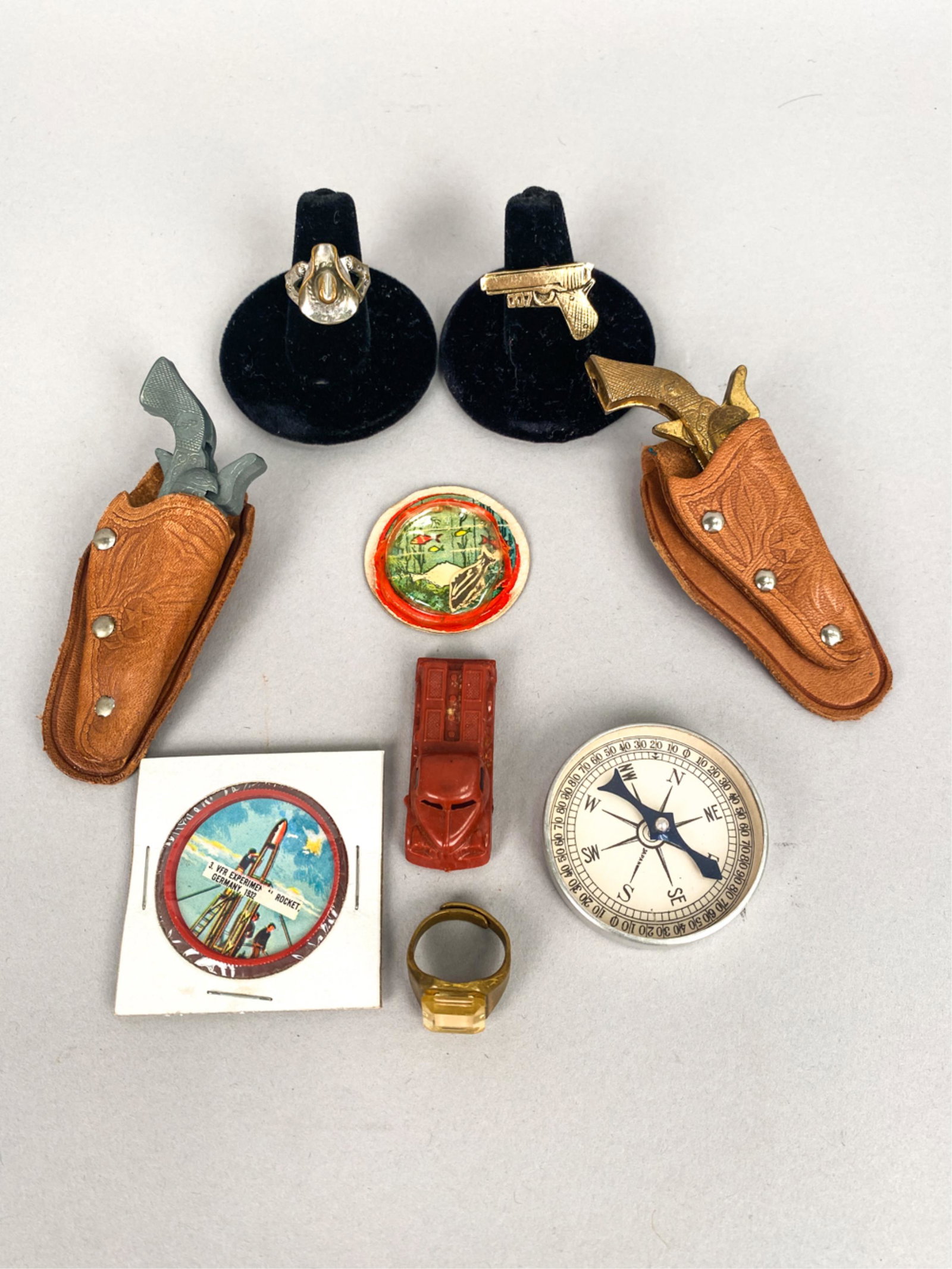 GROUPING OF COLLECTIBLES: Including (3) Rings, (2) Revolvers in holsters, (1) Compass, (1) Gordons potato chips coin, (1) Compass, (CJ & co. Magic puzzle. Dimensions: (Revolver) H 4" x W 1.5" Condition: Revolvers with heavy ta