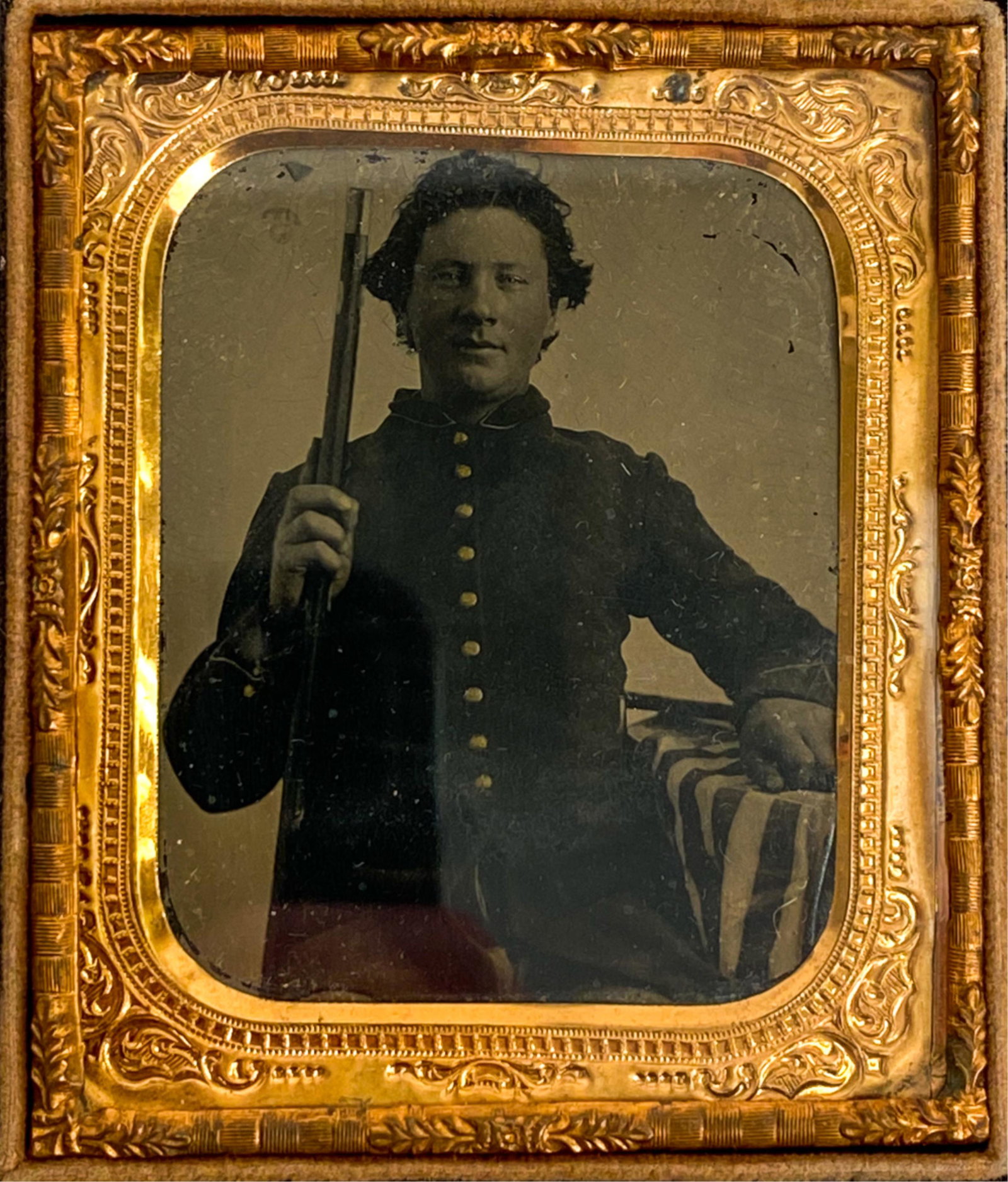 1/6 PLATE TINTYPE JAUNTY YOUNG SOLDIER: Seated private with rifle at table covered by American flag. No hat and tousled hair. Dimensions: (Case) H 3.75" x W 3.25" Condition: Case missing top. Case with damages. Photo clear, with some surfac