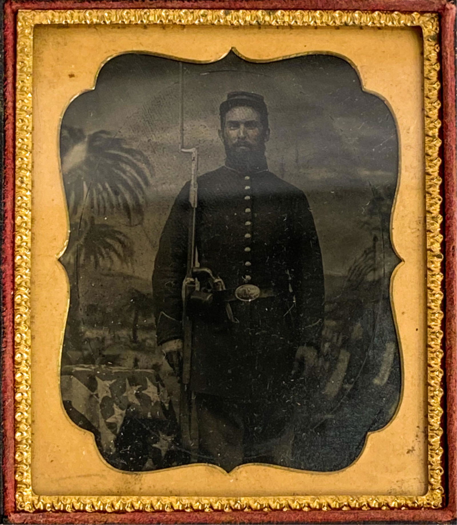 1/6 PLATE TINTYPE 3/4-LENGTH SOLDIER: Union soldier, standing with rifle with affixed bayonet, pistol in belt. In front of a backdrop with palm trees and American flags. Dimensions: (Case) H 3.75" x W 3.25" Condition: Image clear. Somewha