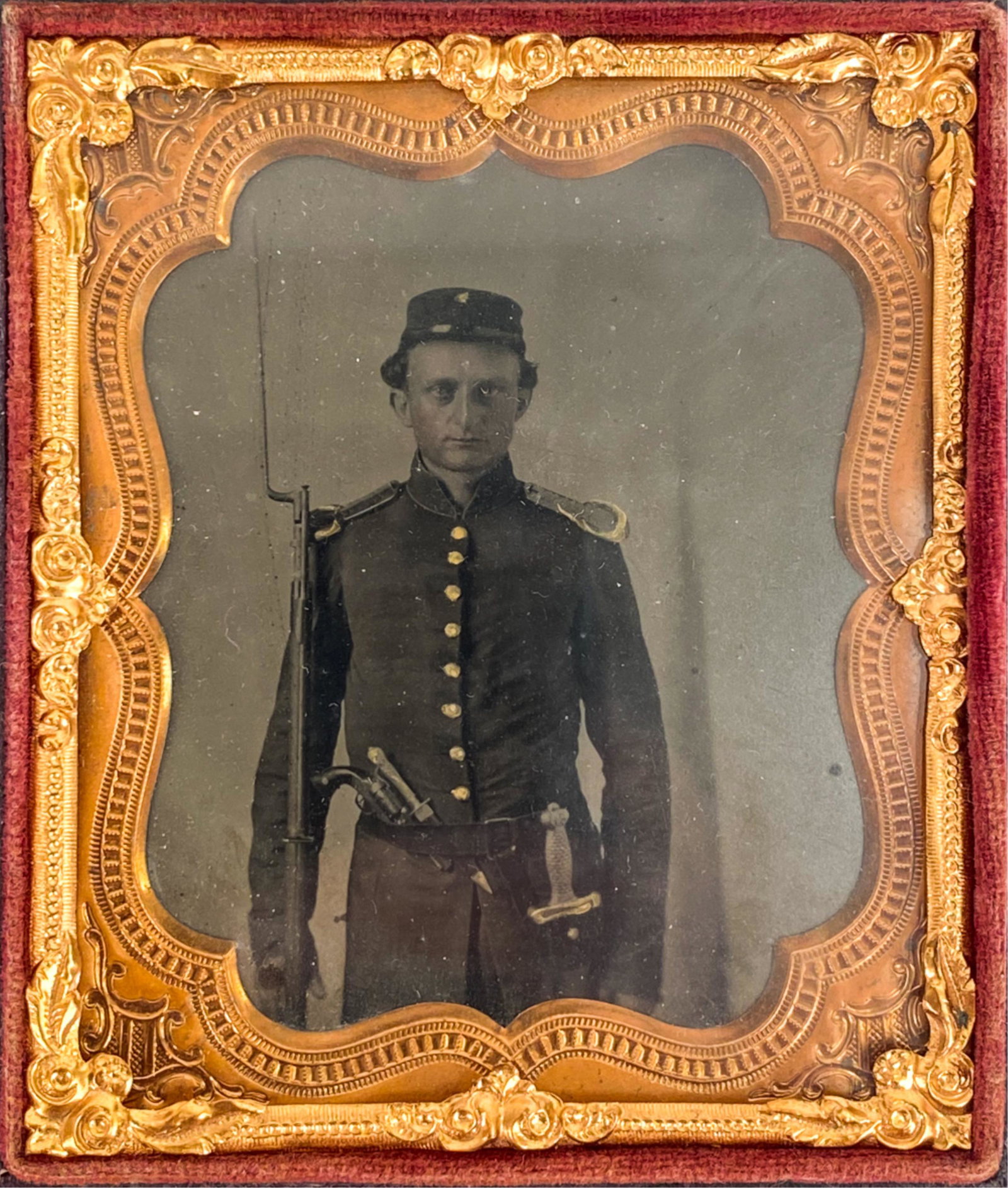 1/6 PLATE TINTYPE FULLY ARMED YOUNG SOLDIER: Earnest young man, staring straight ahead. Triple armed, holding rifle with affixed bayonet. Pistol and dagger in belt, sword at side. Case with bird and grapes. Dimensions: (Case) H 3.75" x W 3.25" C