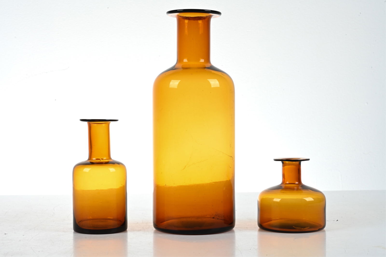 (3) MANNER OF OTTO BRAUER AMBER BLOWN GLASS VASES: (3) Scandinavian Modern studio glass bottles or vases, in the manner of Otto Brauer for Holmegaard. Dimensions: (Largest) H 12.5" x Dia. 5" Condition: No issues to note; no signs of chips, cracks, or