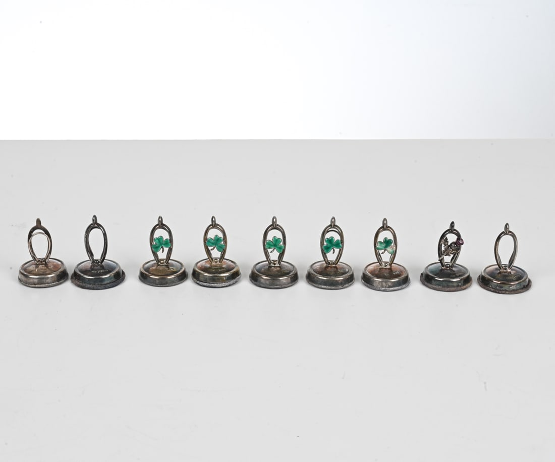 (9) ENGLISH STERLING HORSESHOE PLACECARD HOLDERS: First half of the 20th Century. (5) With enamel clovers, (1) with oak branch. Dimension: H 2" x Dia. 1.25" Condition: A few small dings and bends to metal. Surface tarnish. One base detached; losses t