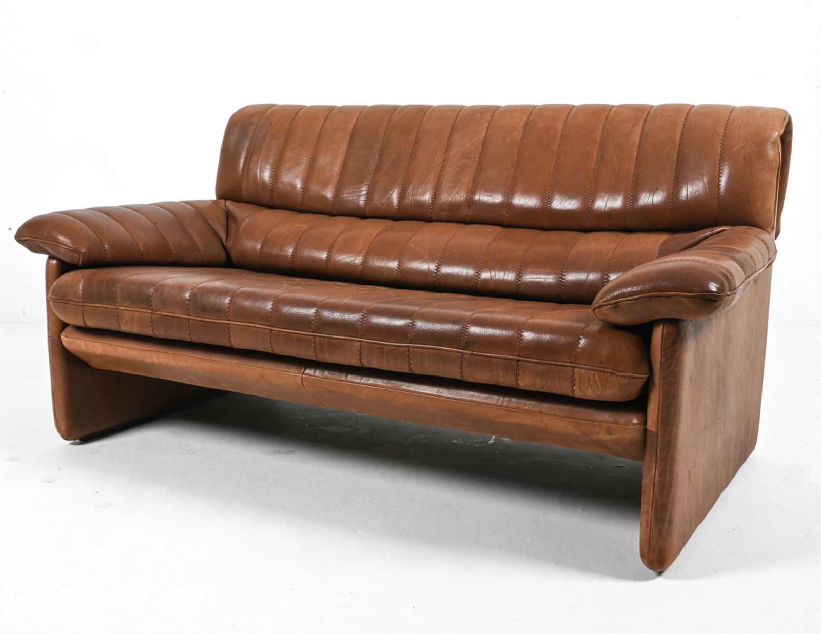 DE SEDE DS-86 BUFFALO LEATHER TWO-SEAT SOFA: De Sede Model DS-86 two-seat sofa in channel-stitched buffalo leather. Switzerland, c. 1970's. Dimensions: H 30.25" x W 54.25" x D 35" x SH 16". Condition: Scratches and scuffs throughout consistent w