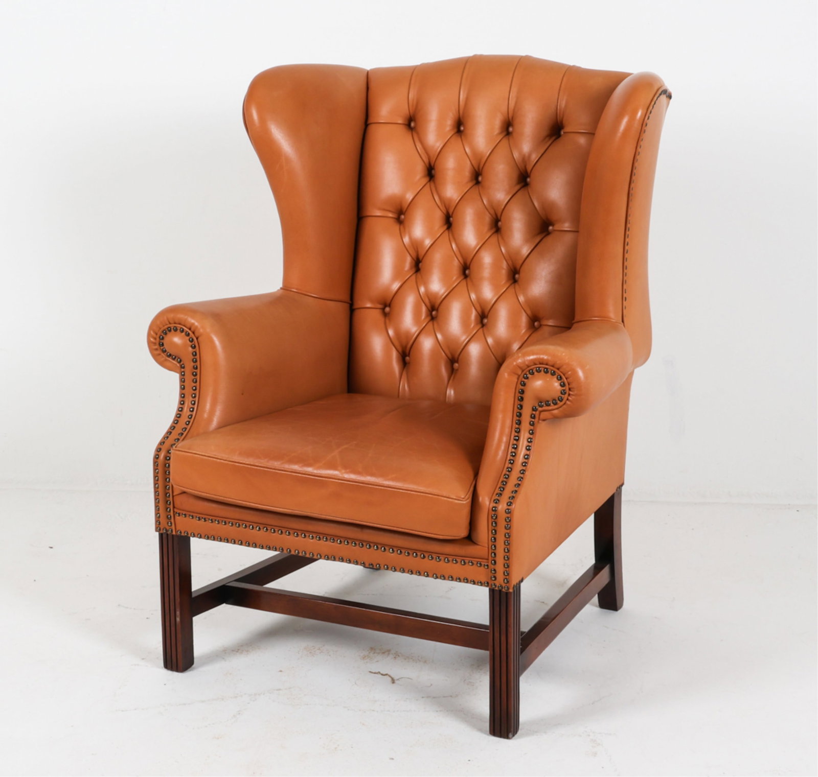 MID-CENTURY ENGLISH LEATHER WINGBACK ARMCHAIR: Tan leather upholstery with brass tacks. Beech legs. Circa 1970's. Dimensions: H 42" x W 35" x D 30" x SH 18". Condition: Shallow gouge on right side, pictured. Some faded areas to leather on arms, ba