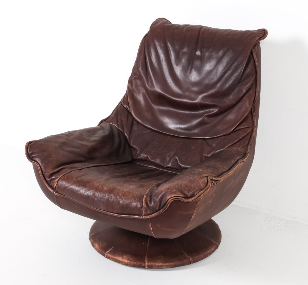 MID-CENTURY DUTCH LEATHER SWIVEL CHAIR C. 1970S: In the manner of De Sede. Brown-tone leather upholstery with leather wrapped swivel base. Dimensions: H 37" x W 33" x D 33.5", SH 18" Condition: Moderate sun fading, creases and scattered blemishes to