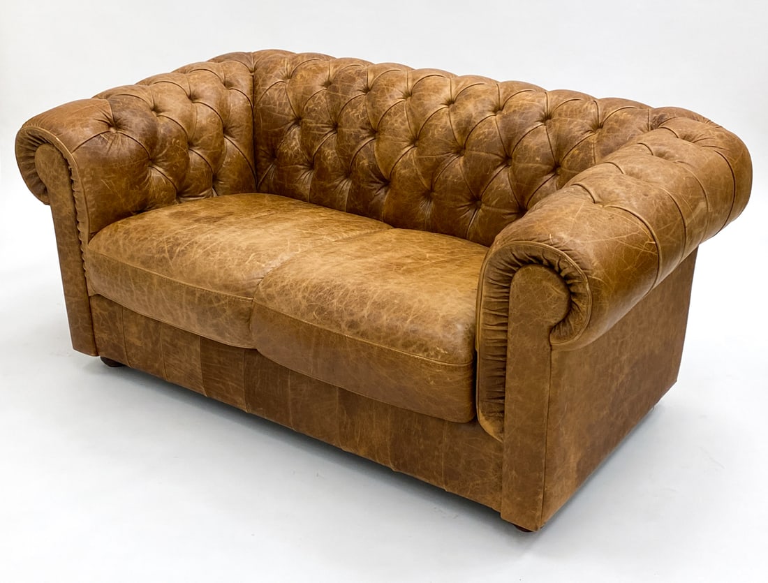 CHESTERFIELD STYLE LEATHER LOVESEAT C. 1960S: Brown tufted leather upholstery. Beech legs. Dimensions: H 27" x W 61" x D 33" x SH 16.5". Condition: Some drying to leather, fading and discoloration to seat and arms. Tobacco odor. Wear consistent w