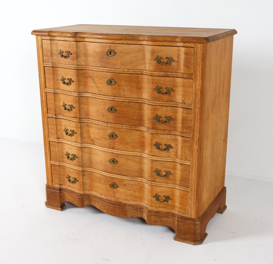 DANISH OAK CHEST OF DRAWERS C. 1970S: Key included. Oak chest of drawers with serpentine front. Brass drawer pulls and locks. Dimensions: H 39.25" x W 37.25" x D 18". Conditions: Wear consistent with age and use. Few water marks and sun f