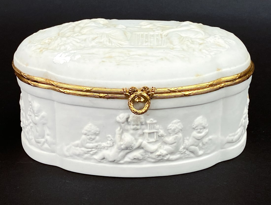 COQUET FRENCH LIMOGES PORCELAIN DRESSER BOX: Coquet bisque white porcelain box with carved Putti details and saints, with gilded details. Marked on underside. 20th century. Dimensions: H 3.5" x W 7.25" x D 6" Condition: Patina to gilded area and