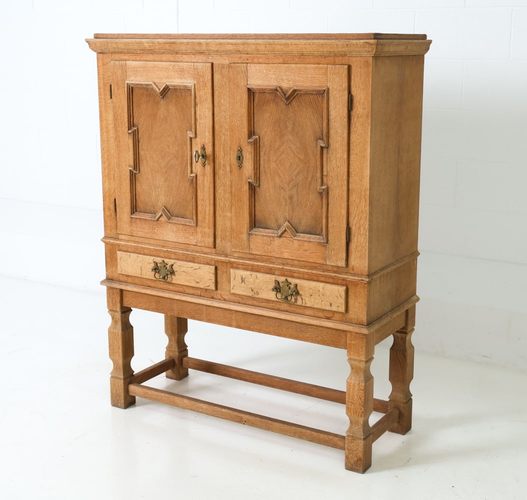 KJAERNULF STYLE DANISH OAK CABINET C. 1960S: One key included. Paneled doors with carved detailing and two drawers with brass pulls. Raised on legs joined by stretchers. Interior fitted with floral paper lining. Denmark, circa 1960s.