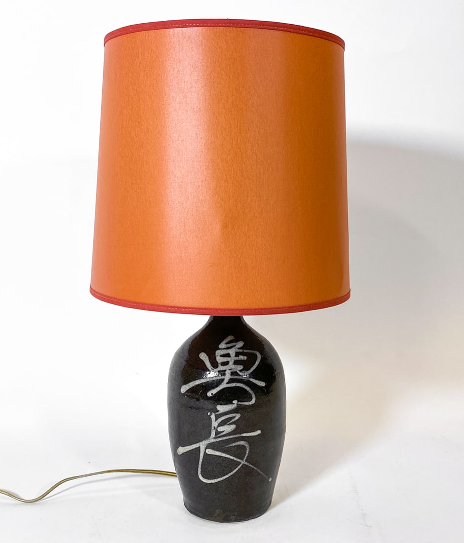 JAPANESE SAKE BOTTLE CERAMIC TABLE LAMP C. 1970S: Ceramic sake bottle converted to table lamp. Dimensions: w/ shade H 20" x Dia. 10.5". Bottle H 11.5". Condition: Wiring untested, cord cut. Wear consistent with age and use.