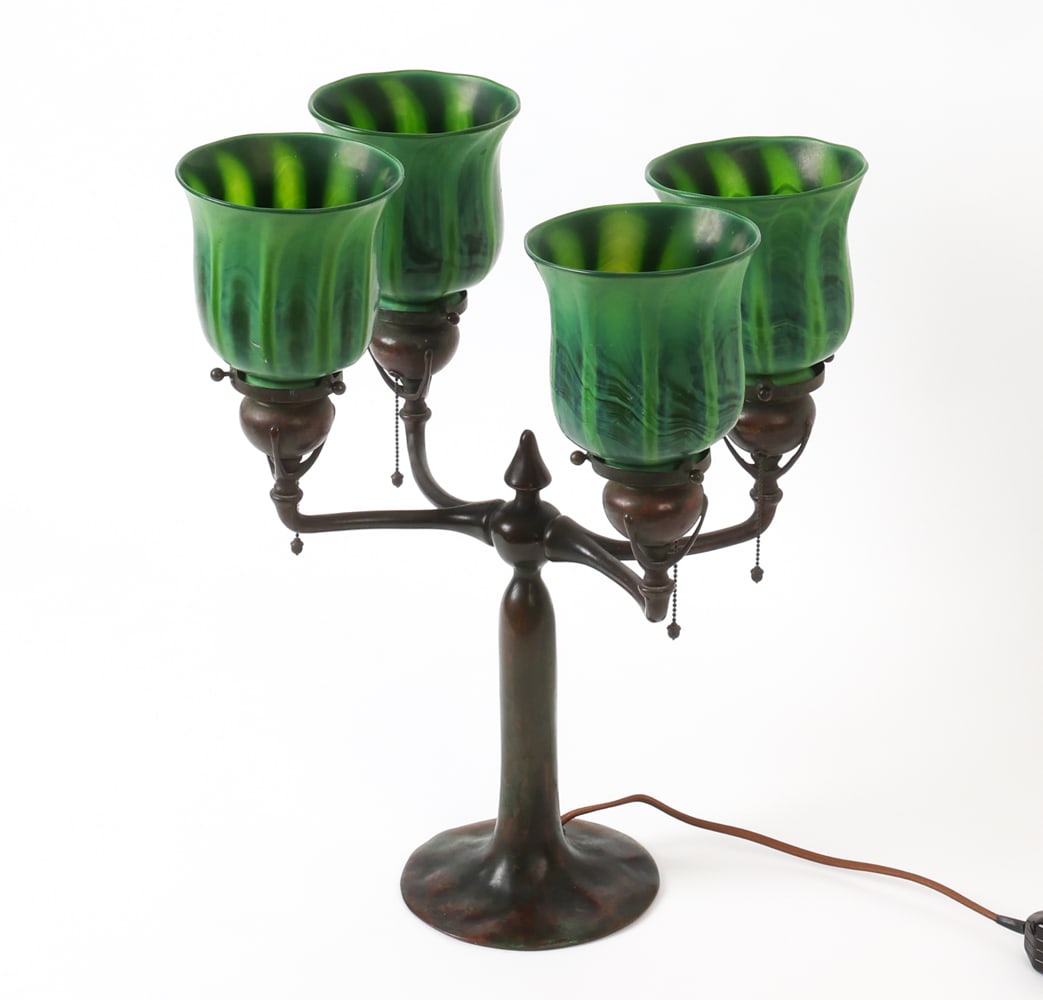 TIFFANY STUDIOS FAVRILE GLASS & BRONZE TABLE LAMP: American Art Nouveau candelabrum table lamp with four-post structure. Favrile glass shades in flared tulip-form and in tones of emerald and darker green, and patinated bronze standard. Pull chains fro