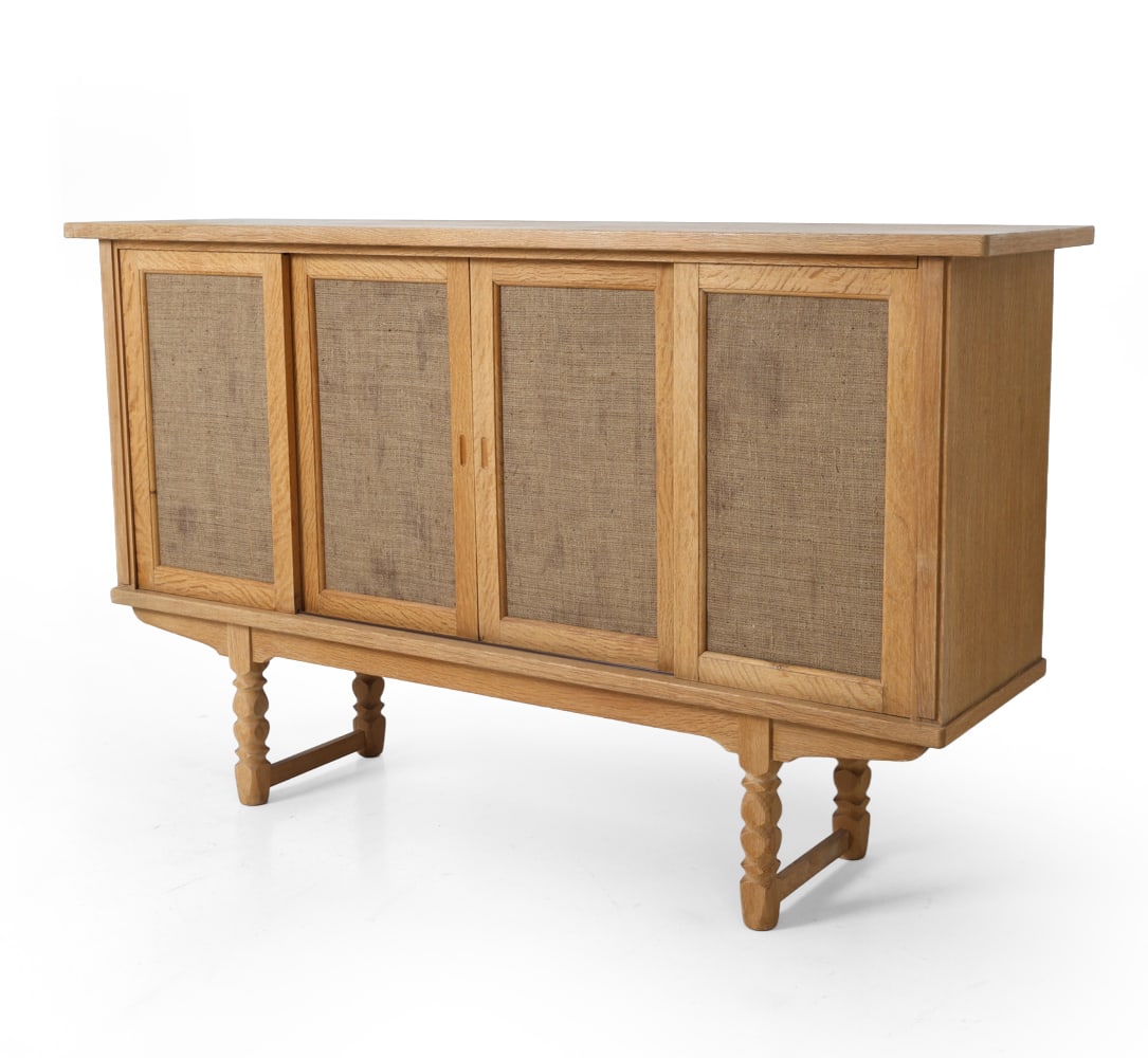 KJAERNULF STYLE DANISH OAK SIDEBOARD 1970S: Oak sideboard with sliding cabinet doors. Woven fabric inset panels to cabinet doors. Removable shelves. Turned legs. Denmark, circa 1970s. Dimensions: H 46" x W 76.5" x D 19.25". Condition: Few water
