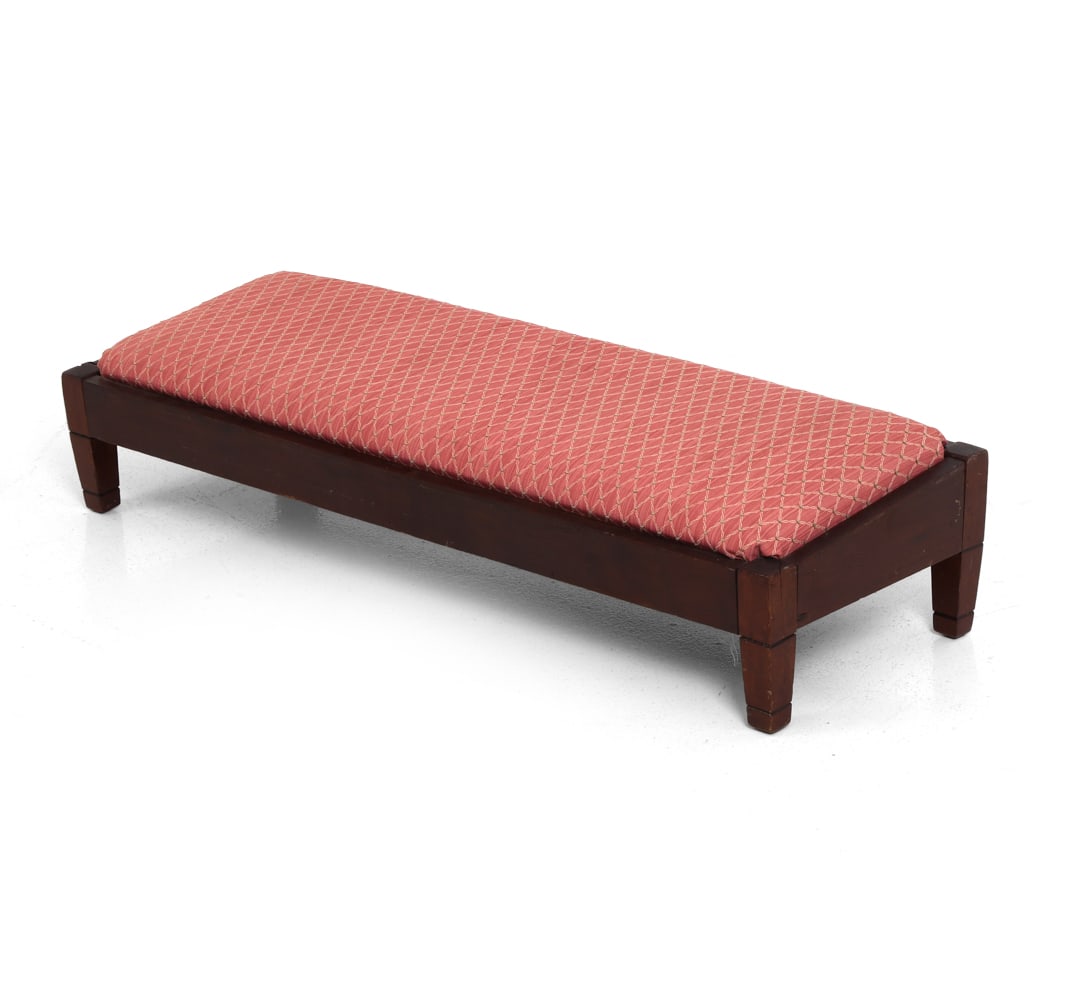 ANTIQUE ANGLED FIRE FENDER FOOTSTOOL: Long upholstered oak footstool. Dimensions: H 8" x W 36.5" x D 12.5". Condition: Light fading to fabric. Scuffs and nicks to frame. Splits and separation to wood. Has been restained and cushion