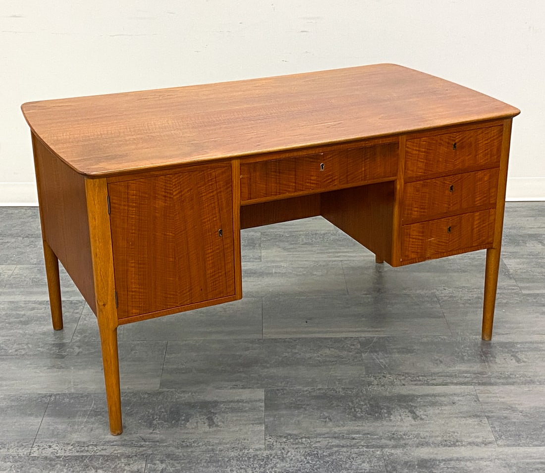 MID-CENTURY DANISH TEAK WRITING DESK C. 1960S: Four drawers, a door and a writing surface desk. Key included. Denmark, circa 1960s. Dimensions: H 29.5" x W 52" x D 29.5" Condition: Wear consistent with age and use. Scuffs, fading discoloration to