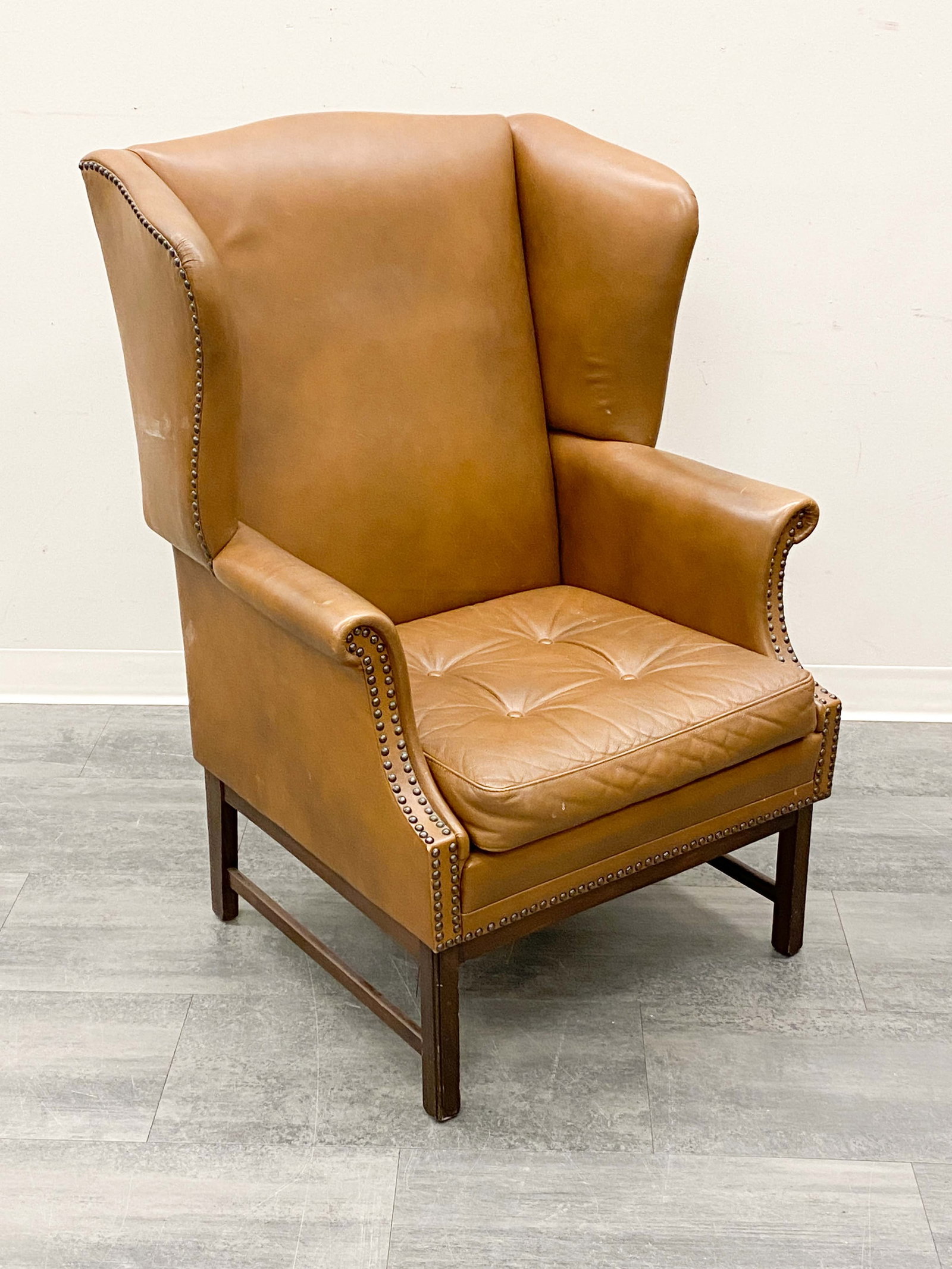 LEATHER WINGBACK LOUNGE CHAIR C. 1950S: Studded detail and leather upholstery. Dimensions: H 43" x W 30.5" x D 26" x SH 16" Condition: Wear consistent with age and use. Minor white soiling marks to sides. Fading, some discoloration, scuffs.