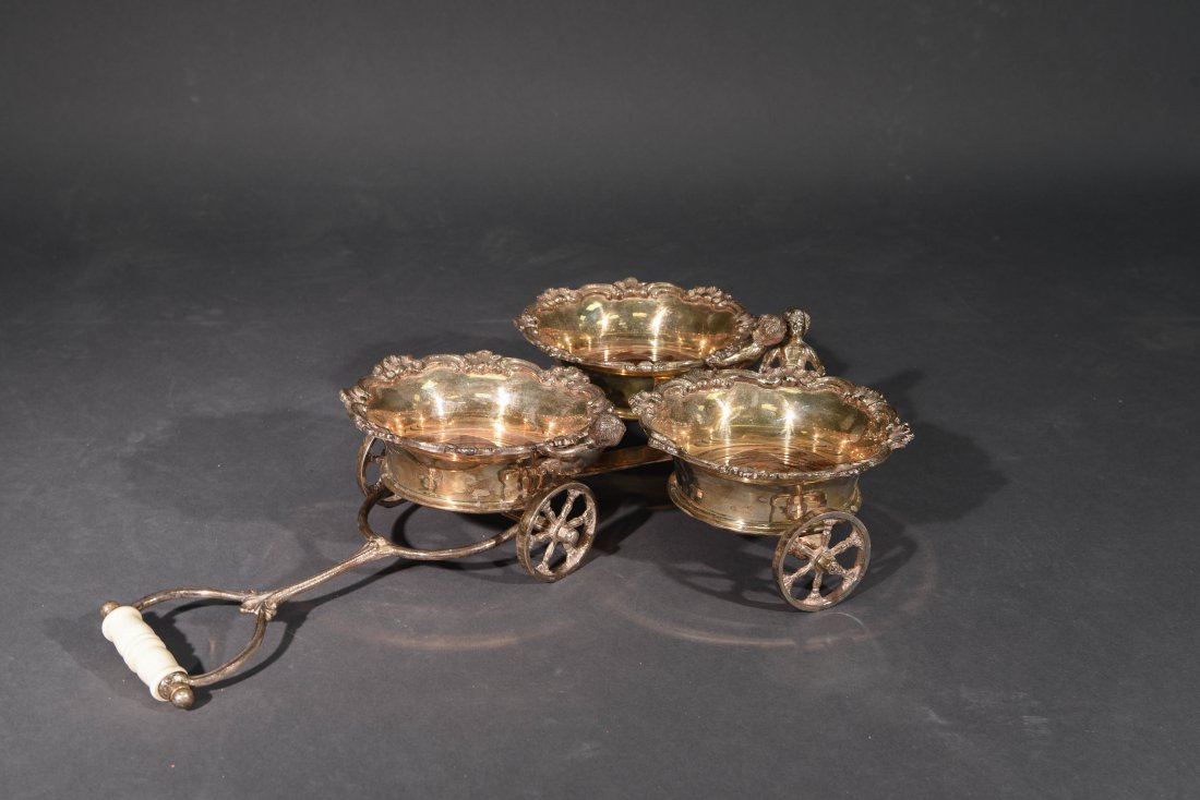 SILVER PLATED WINE TROLLY: Victorian Silver Plate Wine Trolley Coaster, triple trolley form with applied scroll and shell border and putti Dimensions: H 5.5" x W 14" x D 20"