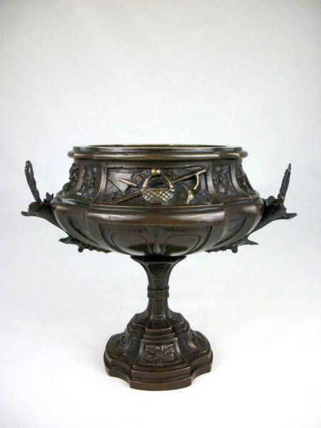 Miniature Bronze Urn, Circa 1900: With maiden-form handles each surmounted by an acanthus headdress, portions of the body decorated with various tools and weapons. Probably French, circa 1900. H. 5 inches.Provenance: Ex-collection Joh