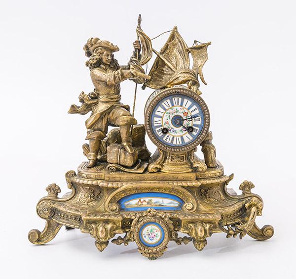 French Gilded Figural Mantel Clock: Antique French mantle clock with pirate figure holding a flag and gun with embossed garments. Sail pediment above a round clock with porcelain face. Porcelain face decorated blue and gilt border with 