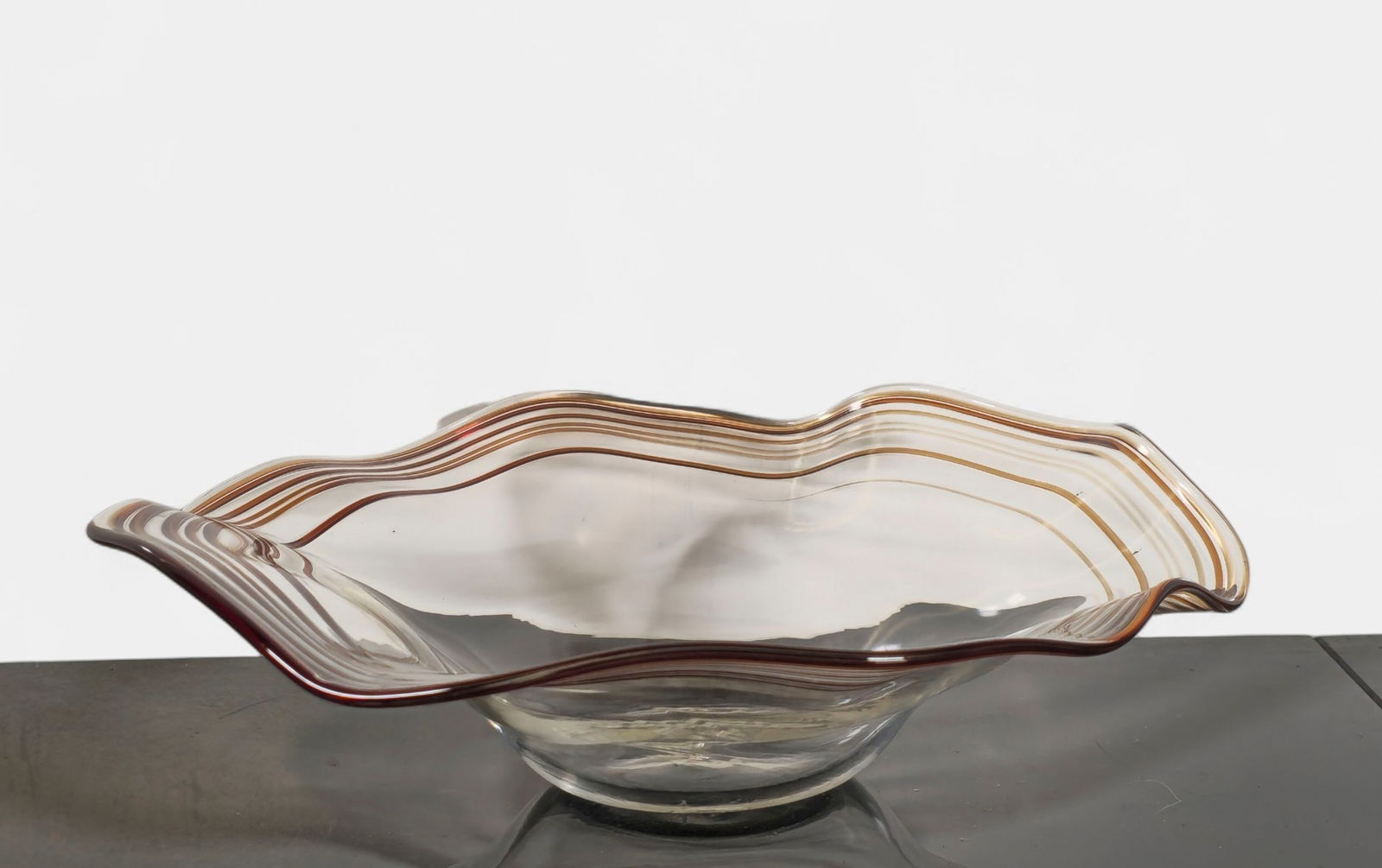 Richard Blenko Hand-Blown Art Glass Bowl: Mid century modern clear art glass bowl with undulating rim and applied amber and cranberry striping, forming a rippling optical effect throughout the form. Signed and numbered to underside “Ric