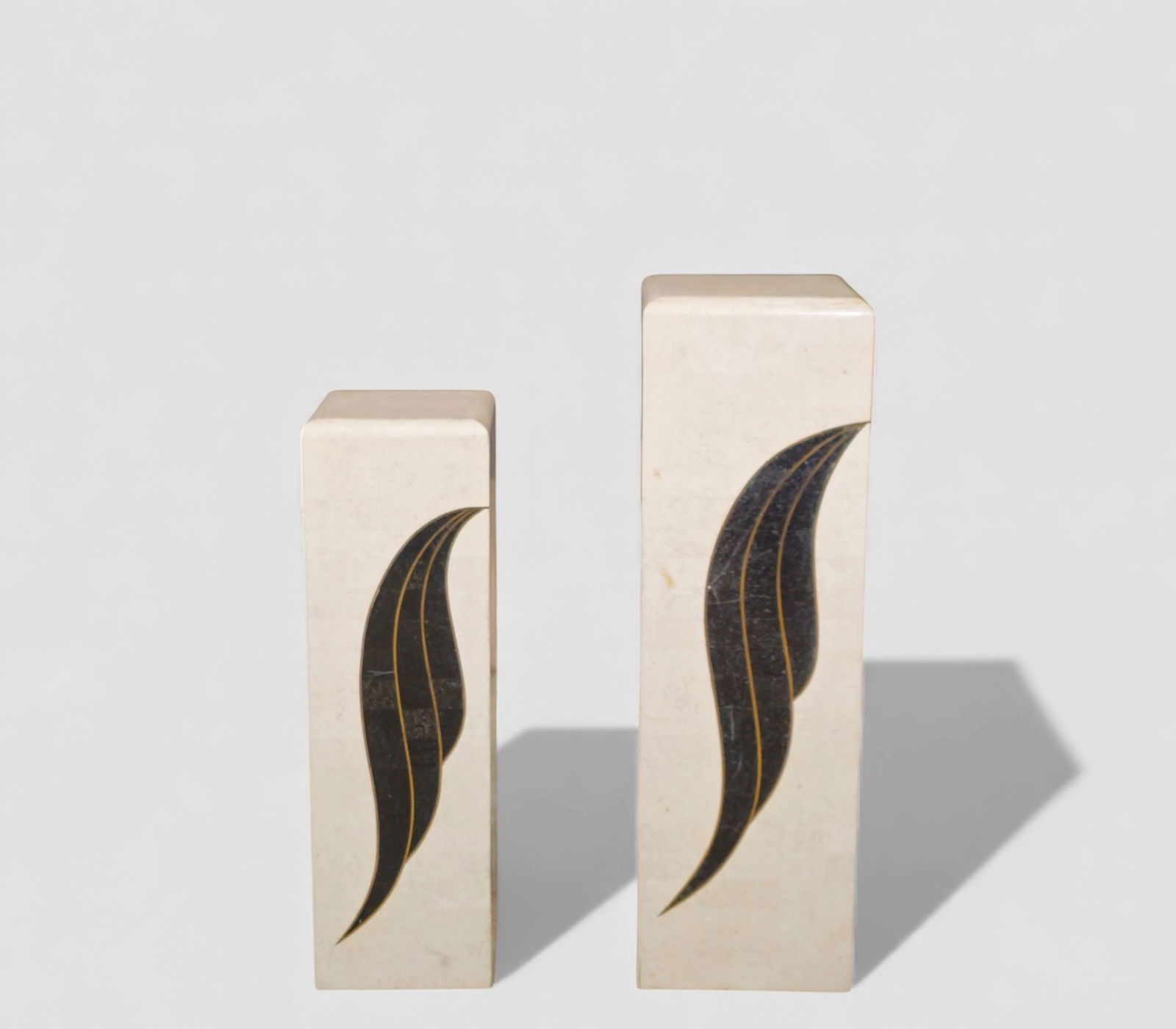 MCM Stone & Brass Pedestals: Two rectangular stone pedestals featuring a minimalist cream-toned surface with stylized black stone leaf inlays outlined in gold. Each column has gently rounded top edges and a smooth honed finish.