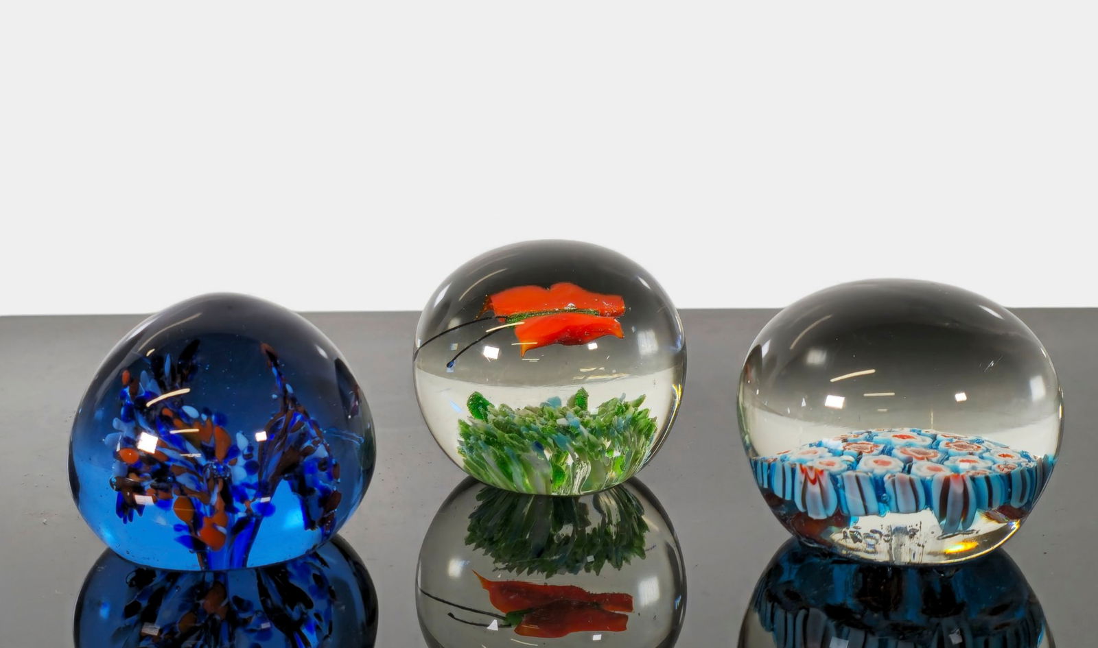 Assembled MCM Art Glass Paperweights: A collection of three mid-century art glass paperweights, each showcasing unique color compositions and internal designs. The group includes: One blue-toned spherical weight with abstract