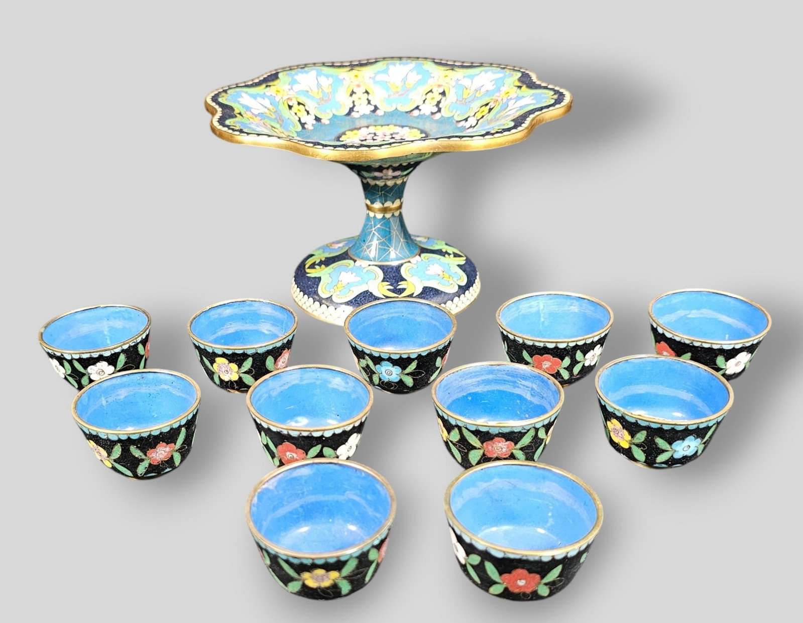 Chinese Cloisonne Enamel Pedestal Compote and Cups: Chinese Cloisonne Enamel Pedestal Compote and Cups. 11 cups. Floral Motif over navy and black ground. 7 1/8w x 7 1/4d x 4 1/8h. Cup 2 dia x 1 1/2h.