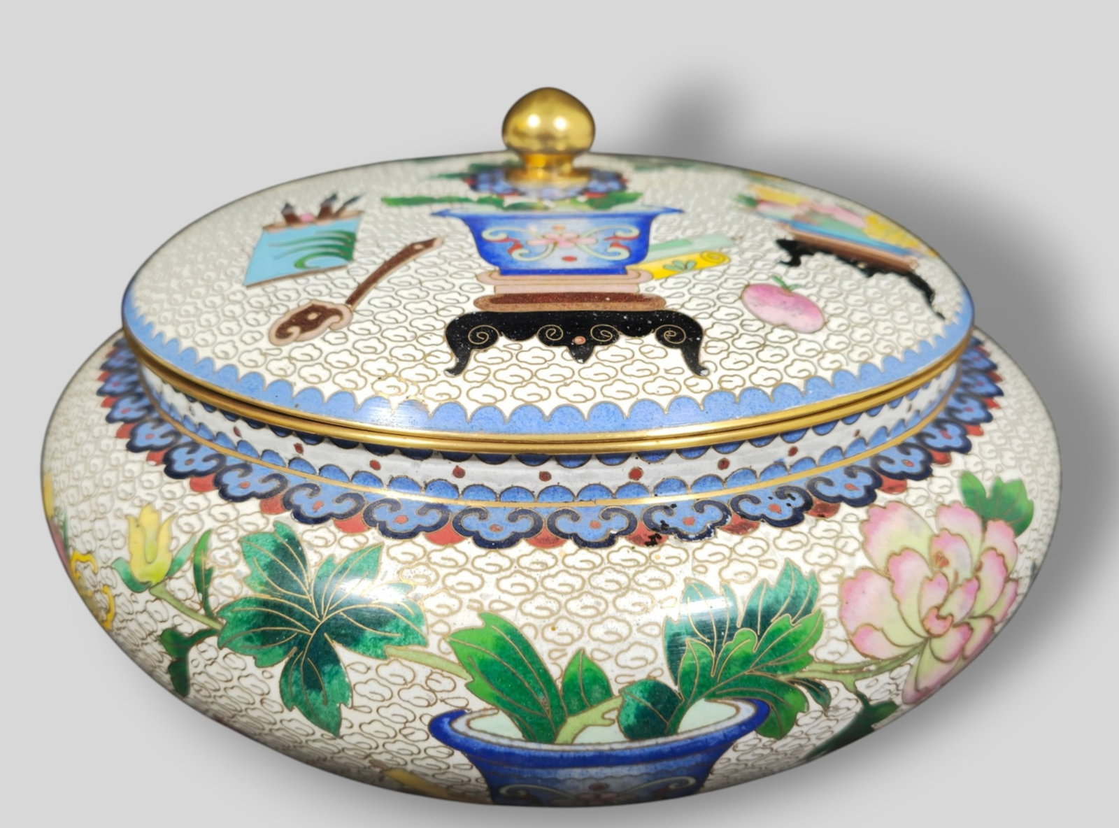 Chinese Cloisonne Enamel Round Lidded Box: Chinese Cloisonne Enamel Round Lidded Box. 20th Century. Decorated with fruit bowl brushes in brush holder and planter on stand. Brass lid handle. 10 dia x 6h.
