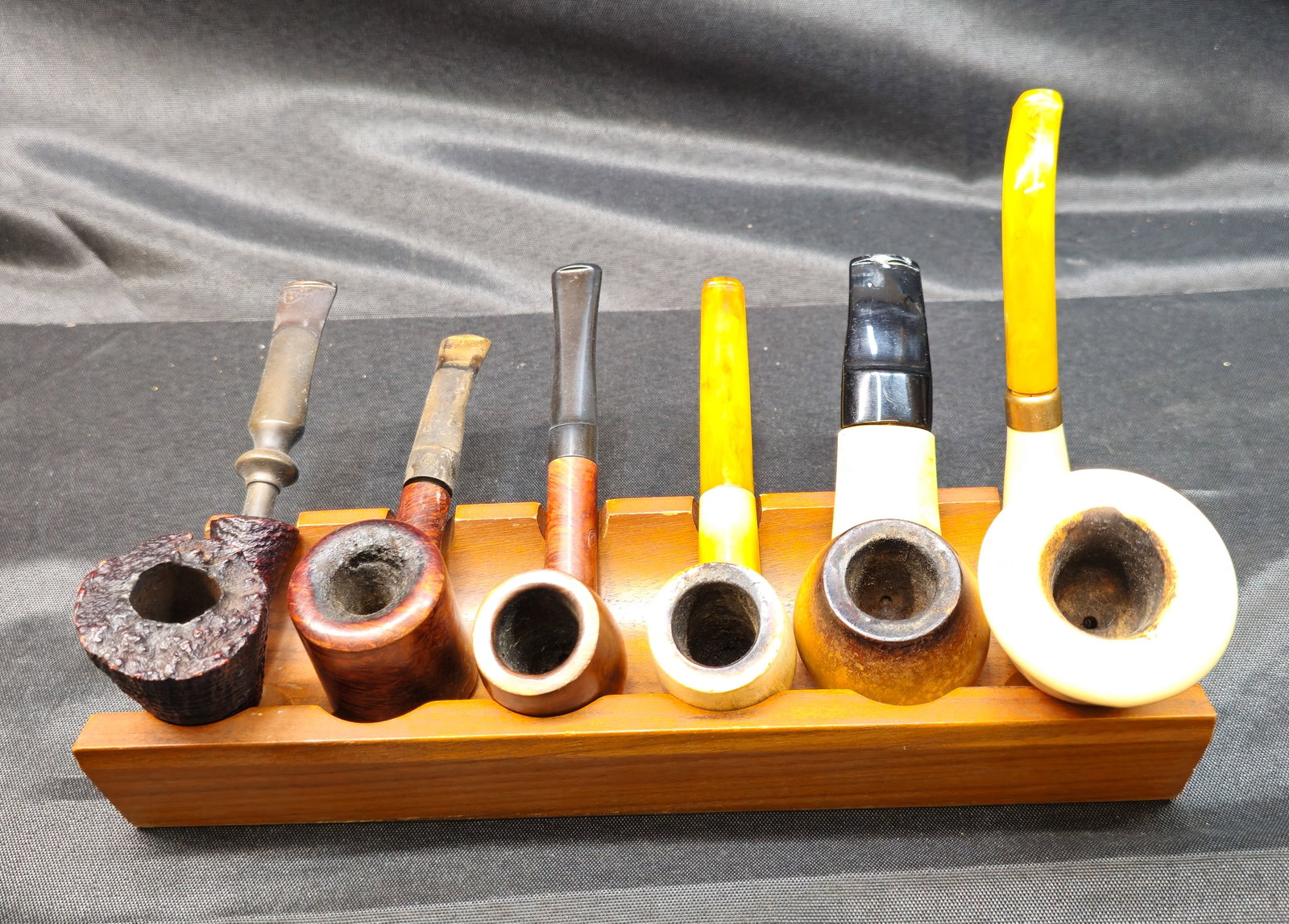 Six Smoking Pipes with Wooden Pipe Rack: Six Smoking Pipes with Wooden Pipe Rack. Assembled collection of six smoking pipes displayed in a fitted wooden pipe rack. The group features an interesting variety of forms, finishes and materials,