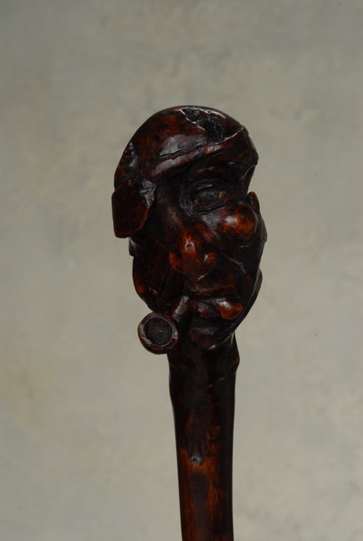 A wood folk art cane of a man with a pipe: An amusing wood folk art cane of a man with a pipe, and his tongue protruding. Made of a single piece of debarked hardwood, it has a carved root handle that is about 3 1/2" high and 2 1/4" at its wide