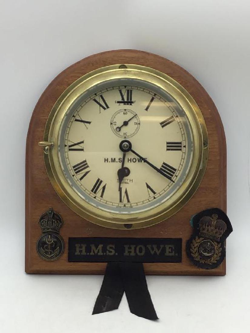 H.M.S. Howe brass Ships Clock: brass clock mounted on wood board with embroidered sash, overall size 10 inches x 10 inches, Made in England, face reads H.M.S. Howe - Smith - Astral. Provenance: From the Estate of Raymond Bentley.