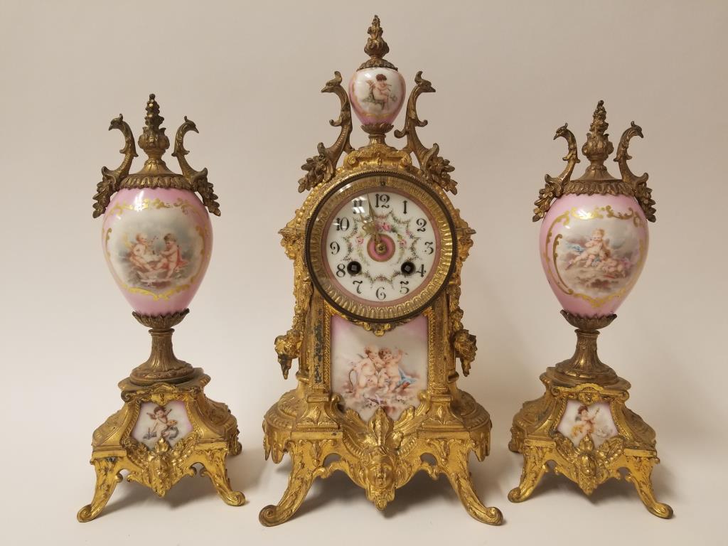 3 piece French Clock Garniture Set: Ornate gilt and hand painted Putti on porcelain. Partial label on base, Clocks and Watches, Philadelphia PA. Clock is 14 inches tall, chimes work, pendulum swings briefly, gilt loss on sides and crack