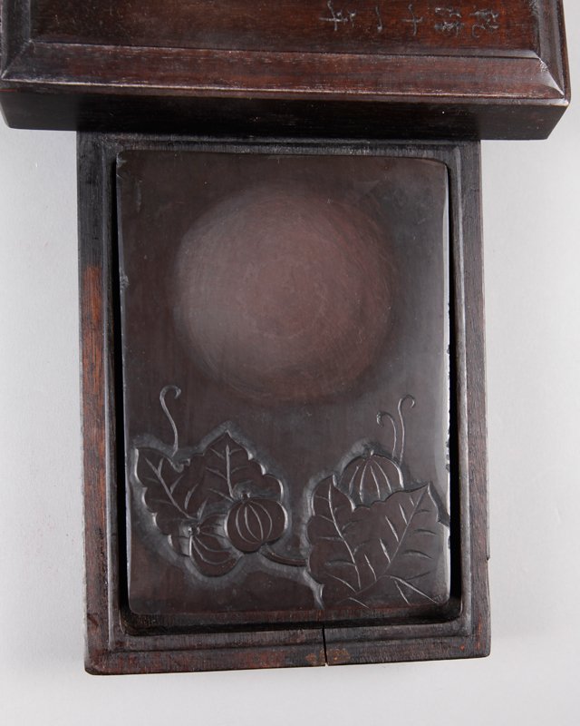 CHINESE GU YUE XUAN INK STONE IN BOX: With leaves and pods to stone, in custom carved wood box with caligraphy and seal mark. Box measures 2" H x 6.5" L x 4.875" W.