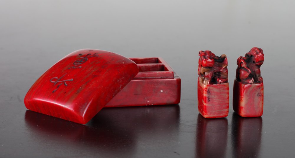 Pair of Chinese Chicken Blood Seals Encased: Two character seals with foo dog carved at top, encased in chicken blood soapstone custom carved box, the lid with caligraphy, the bottom with four character seal mark. Box measures 1" H x 1.875" L x 