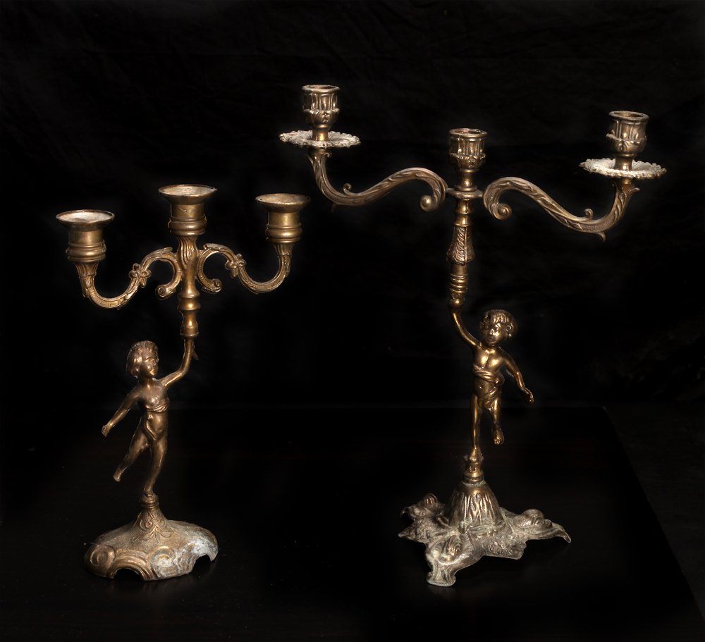 2 Antique Brass Candelabras: Both with three cups, each with similar cherub/child column. Largest measures 14.5" H x 11.75" W. Very good condition with natural patina and wax remnants.