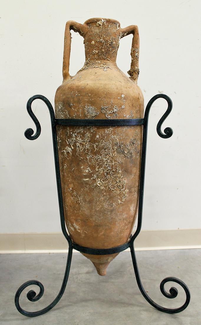 ANCIENT AMPHORA SHIPWRECK OIL VESSEL: Ancient Roman or Greek amphora shipwreck oil or wine vessel with custom metal stand. Hole to bottom side. Measures approximately 36" long and 39" with stand. Discovered off the coast of Elba.