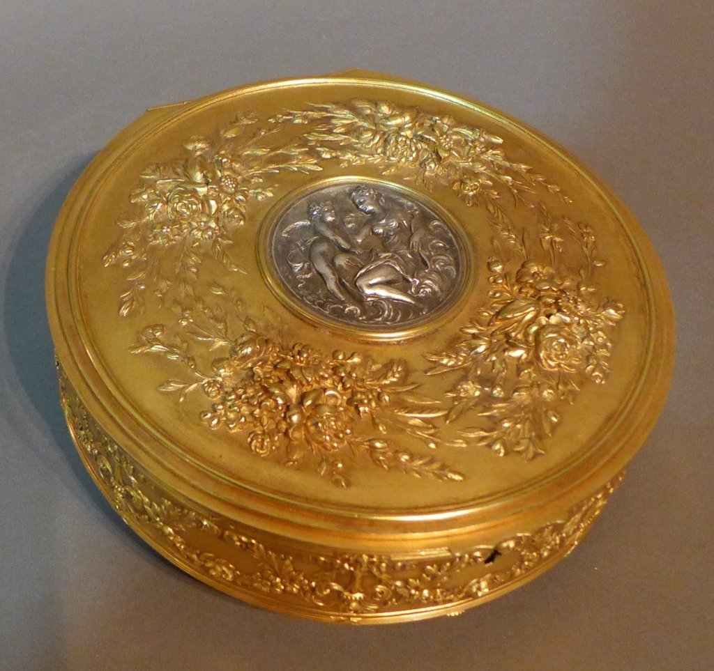 19th Century Silvered and Gilt Box: Dore and silver over bronze circular hinged box on 4 feet. 19th Century, French. Winged putti and female centered plaque on center of lid, surround by floral design. Inside lined with ivory velvet. No