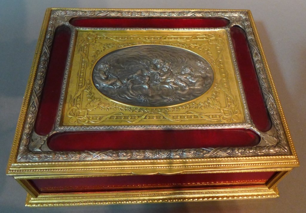 Antique Dore and Silver over Bronze Box: 19th Century French gilt, silvered and red glass hinged box. Figural design of 4 putti on center lid surrounded by deep red glass panels. Inside lining is a luxurious red velvet. Height: 3 1/2", Width