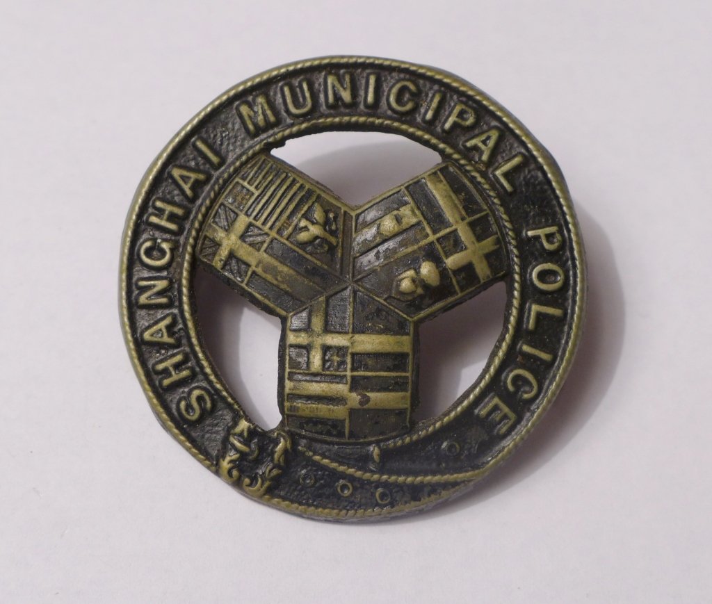 Shanghai Municipal Police Constables Cap Badge: Rare Shanghai Municipal Police Chinese Constables Cap Badge. Featuring pierced front with belt like ring with "Shanghai Municipal Police" sign and flags of all nations in the centre. Imperfections. Di