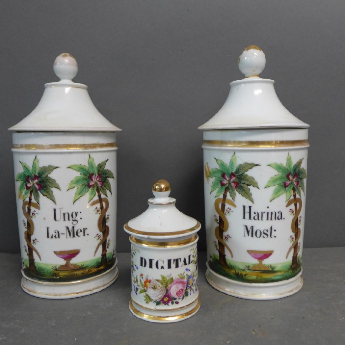 Antique French Porcelain Apothecary Jars: Three hand painted porcelain jars with original lids. Gilt accents. -The two larger containers are decorated with a center chalice between snakes/ serpents slithering up palm trees. Central text reads