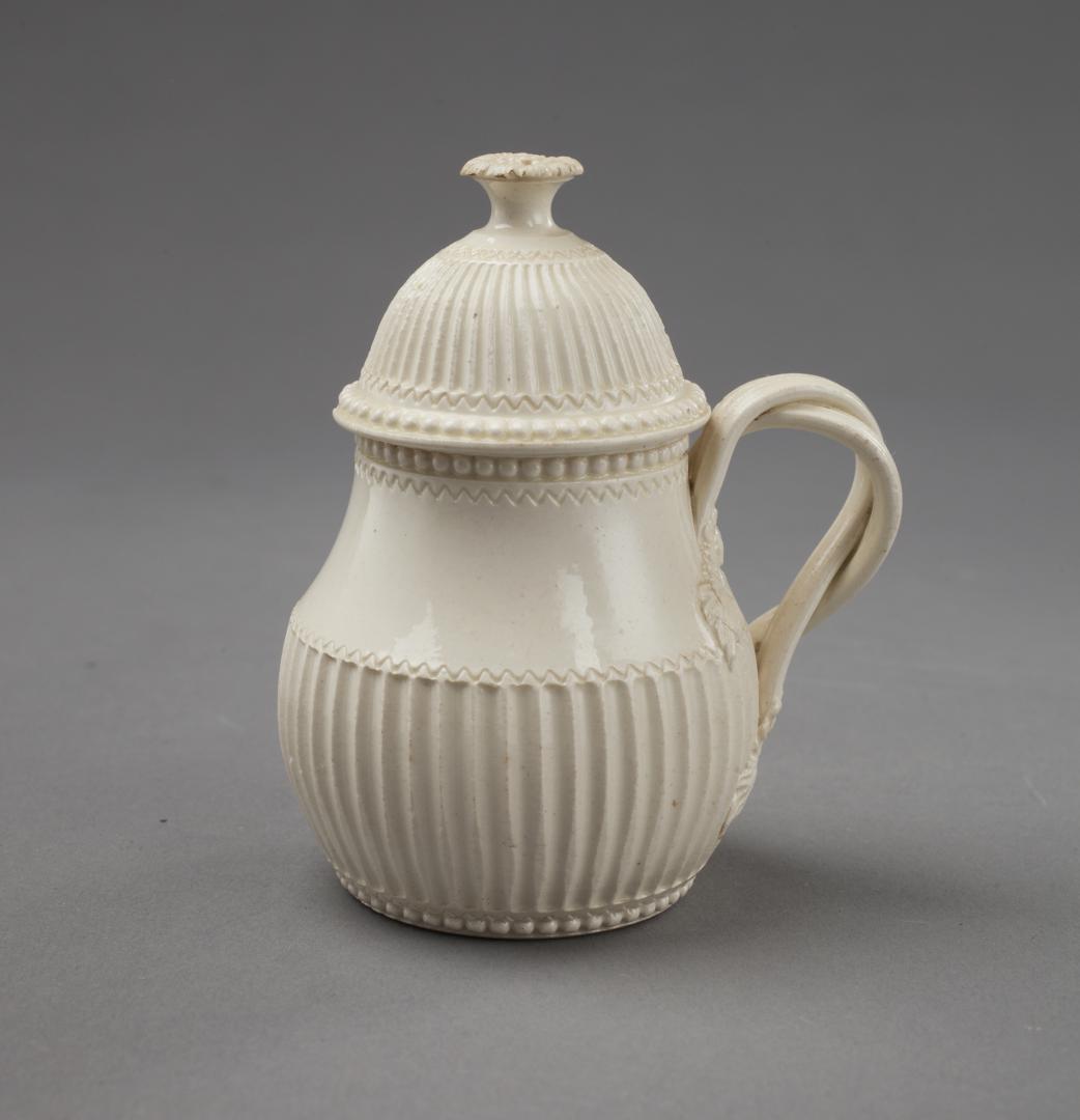 CREAMWARE COVERED MUSTARD CUP WITH FLUTED LOWER HALF: English, circa 1780.Its pear-shaped body with wide band of fluting around its lower body; beading around foot, at rim, and edge of the ribbed cover; button finial; entwined handles. Provenance: A Dist