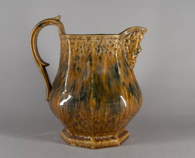 Rockingham Type Stoneware Pitcher: Possibly Bennington, mid 19th century Having paneled sides, a double scroll handle and a mask spout, on a conforming stepped octagonal base, the Rockingham-style glaze with splashes of green. H. 10