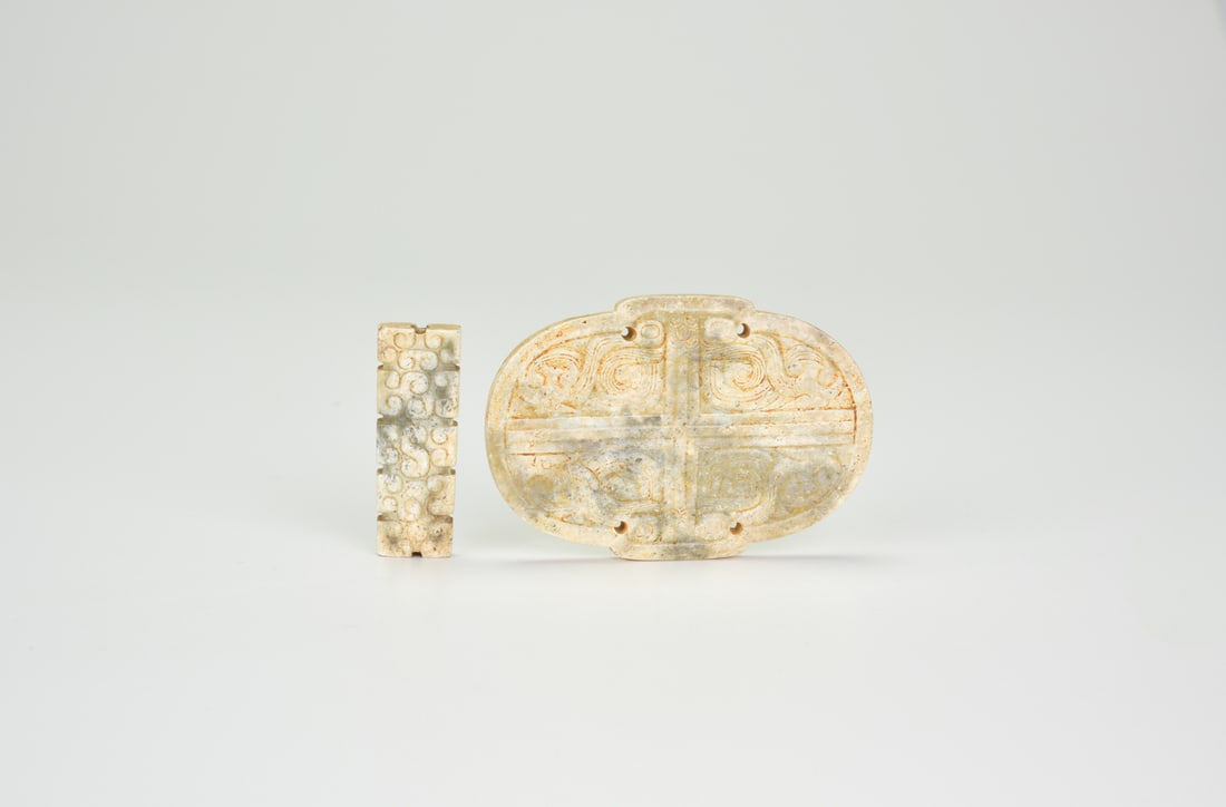 A Carved Celadon And Beige Jade Plaque And A Rectangular Ornament, Shang–Zhou: A Carved Celadon And Beige Jade Plaque And A Rectangular Ornament, Shang–ZhouThe rounded plaque is carved with symmetrical taotie-like scrolls and pierced with four suspension holes, the stone s