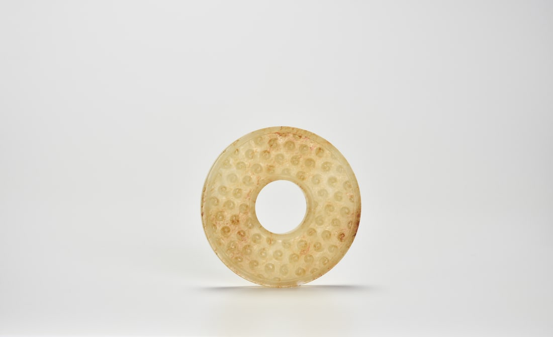 A Jade Bi Disc with Grain Pattern, Han Dynasty (206 BC – AD 220): A Jade Bi Disc with Grain Pattern, Han Dynasty (206 BC – AD 220)Carved from a translucent pale celadon jade with areas of russet and calcified alteration, the disc has a central aperture surroun