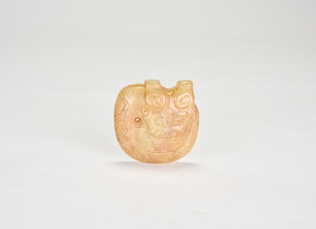 A Jade Pendant Carved as a Coiled Dragon. Shang Dynasty, ca. 13th–11th century BC.: The pendant formed as a compact coiled dragon, The surface carved with incised lines and shallow relief, incorporating geometric triangular motifs and scroll patterns. A perforation at the side.H: 1 3