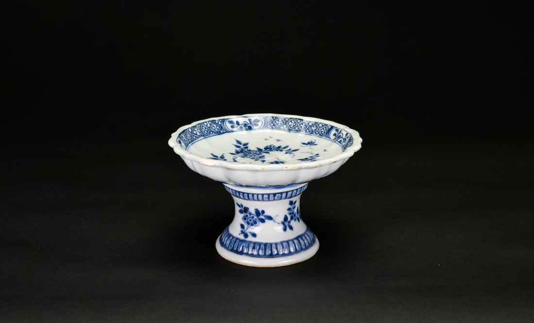 A Blue and White Lobed Stem Dish (Late Qing): The dish is raised on a hollow, flared stem foot, the underside revealing an unglazed footring and recessed interior typical of kiln support. The shallow lobed body with gently scalloped rim is painte