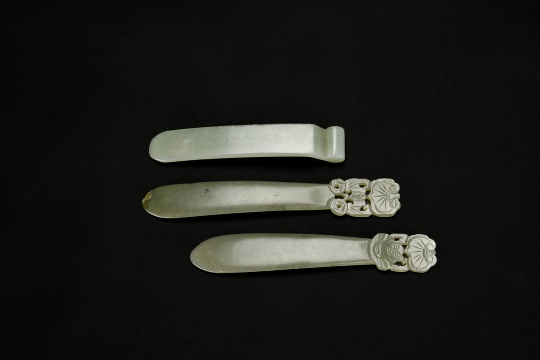 Three Celadon Jade Hairpins Qing Dynasty or Later: Carved from pale celadon jade. Two are surmounted by openwork and relief-carved terminals depicting floral motifs and a smiling figure beneath a stylized leaf, while the third is of plain form with a 