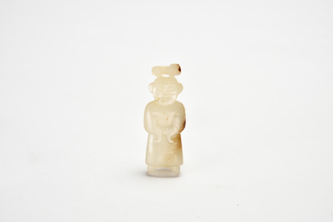 A Jade Figure Of A Standing Attendant, Warring States Period: The figure is carved standing upright with hands held before the chest beneath long sleeves defined by parallel incised lines. The face is softly modeled with almond-shaped eyes and a small mouth bene
