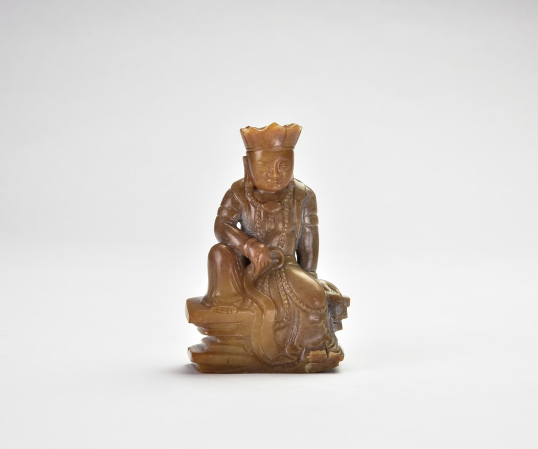 Ming Dynasty Jade Figure of a Seated Bodhisattva: The figure is carved seated in a relaxed pose atop a waisted pedestal base, wearing long flowing robes and an elaborate crown. The serene face is framed by pendulous earlobes, with the right hand rest