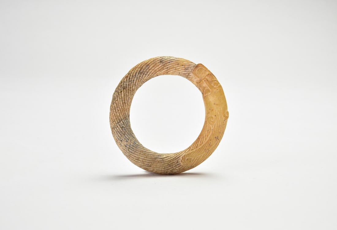 A Chinese Archaic Jade Dragon-Form Ring, Zhou Dynasty: The ring is carved from pale celadon jade, formed as a coiled dragon with the head grasping the tail. The body is finely incised with dense diagonal striations and scroll motifs, the surface with