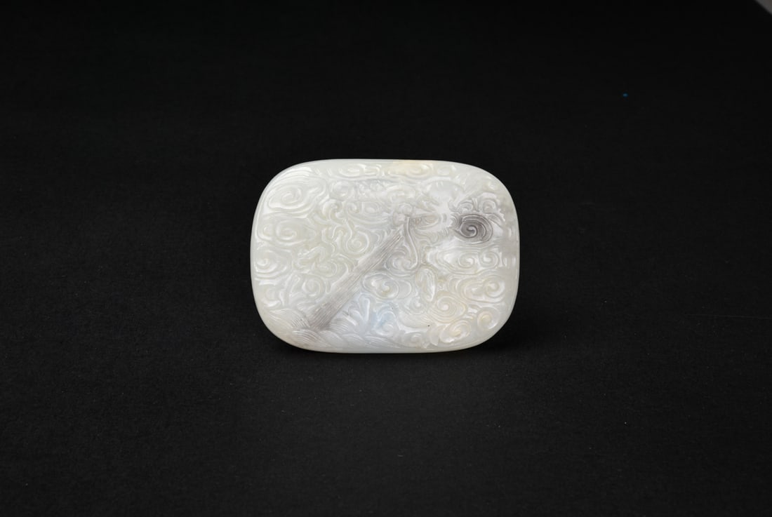 A White Jade Qin and Auspicious Clouds Belt Buckle: Finely carved from a pale celadon-white jade with subtle natural gray inclusions, the belt buckle is decorated in low relief with a qin amidst dense swirling ruyi-shaped clouds, symbolizing scholarly